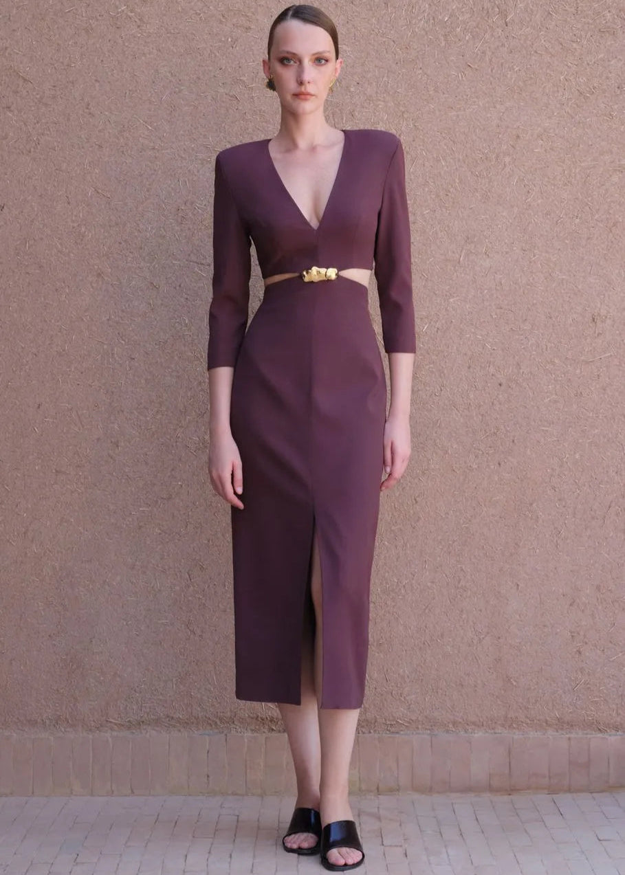 Woman wearing a burgundy dress with a gold belt against a beige wall.