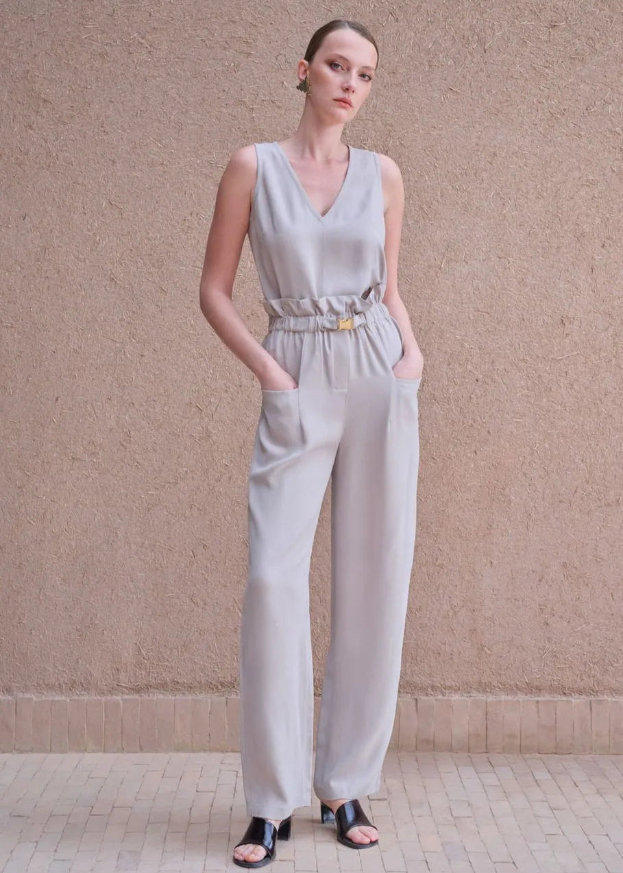 Woman wearing a light gray sleeveless jumpsuit with a belt against a beige wall.