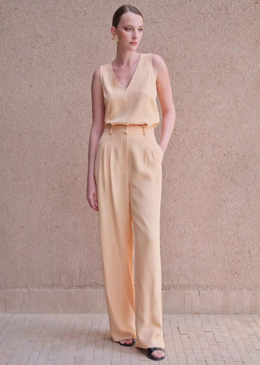 Woman wearing a beige sleeveless top and pants against a plain wall.