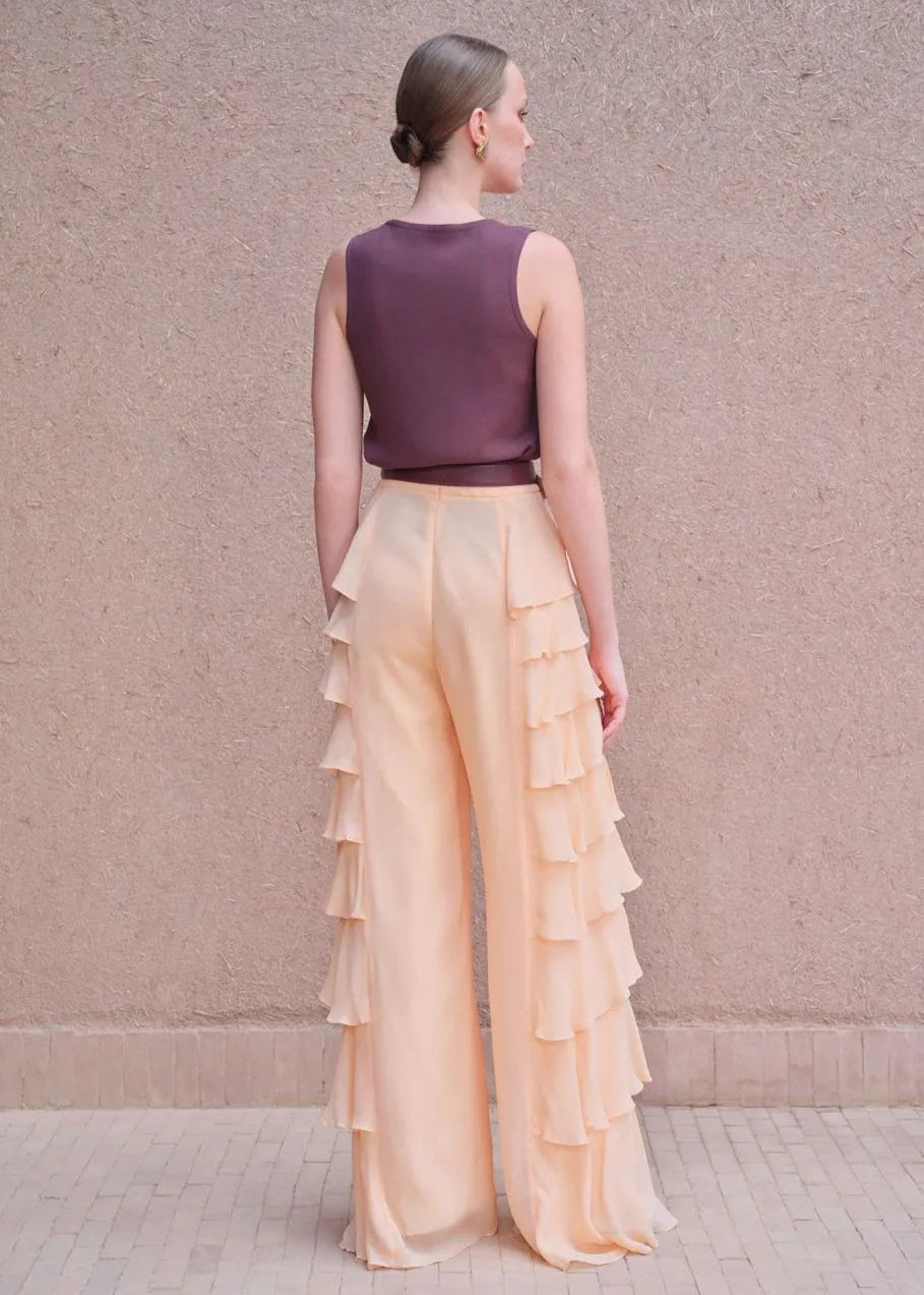 Woman wearing a purple top and beige ruffled pants against a beige wall.