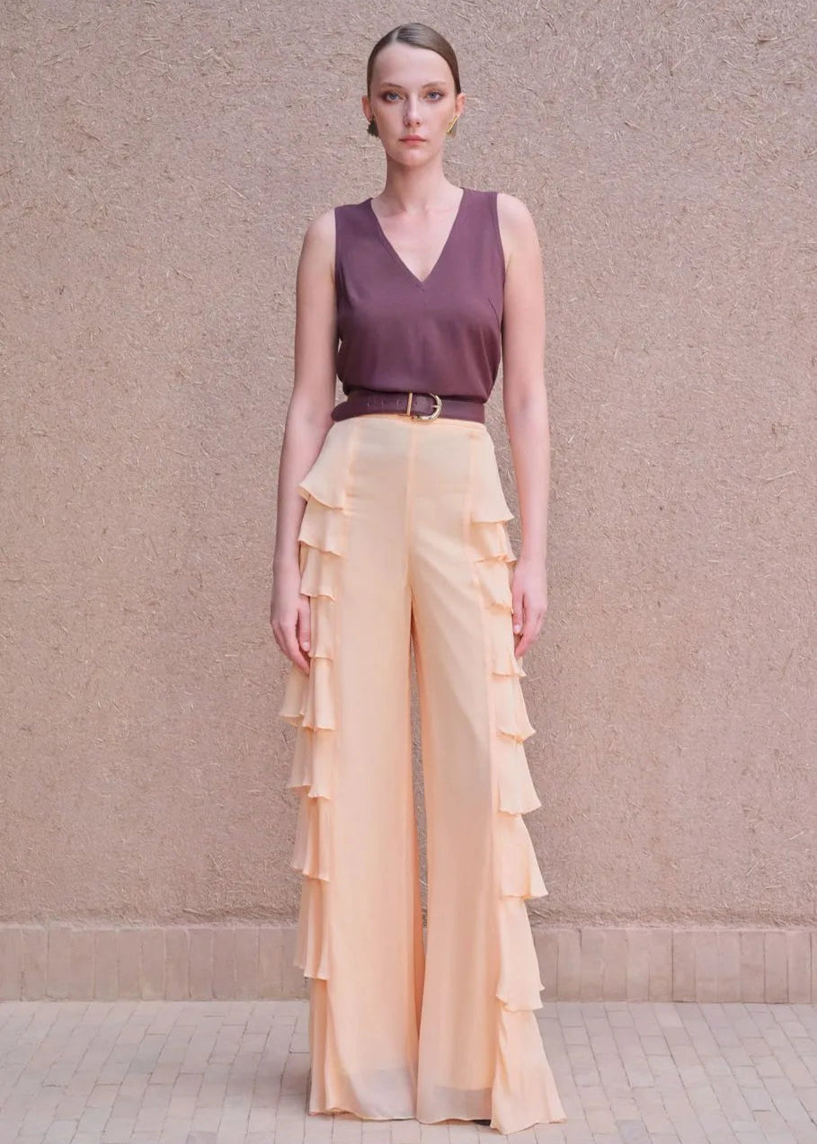 Woman wearing a purple top and peach ruffled pants against a beige wall.