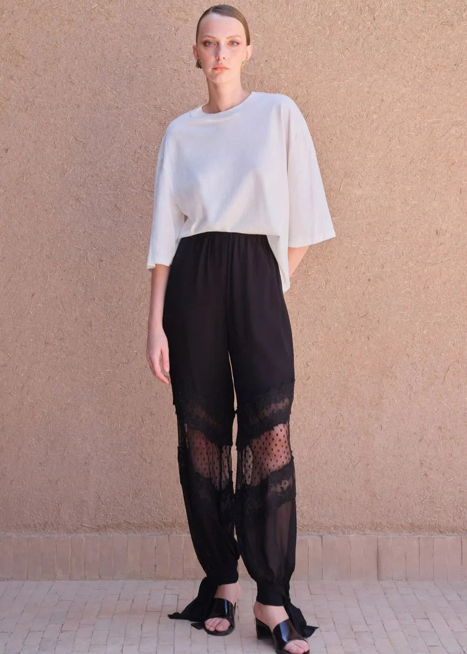 Person wearing a white top and black pants with lace details against a beige wall.