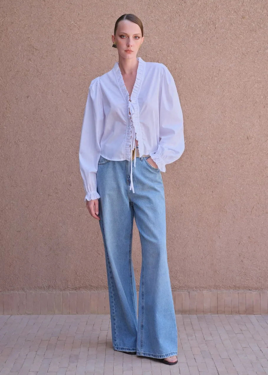 Woman wearing a white blouse and light blue jeans against a beige wall.