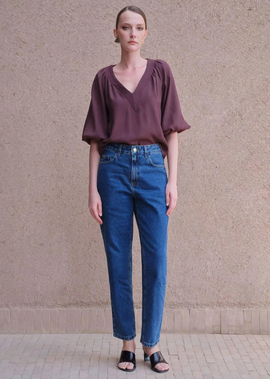 Woman wearing a purple blouse and blue jeans standing against a beige wall.