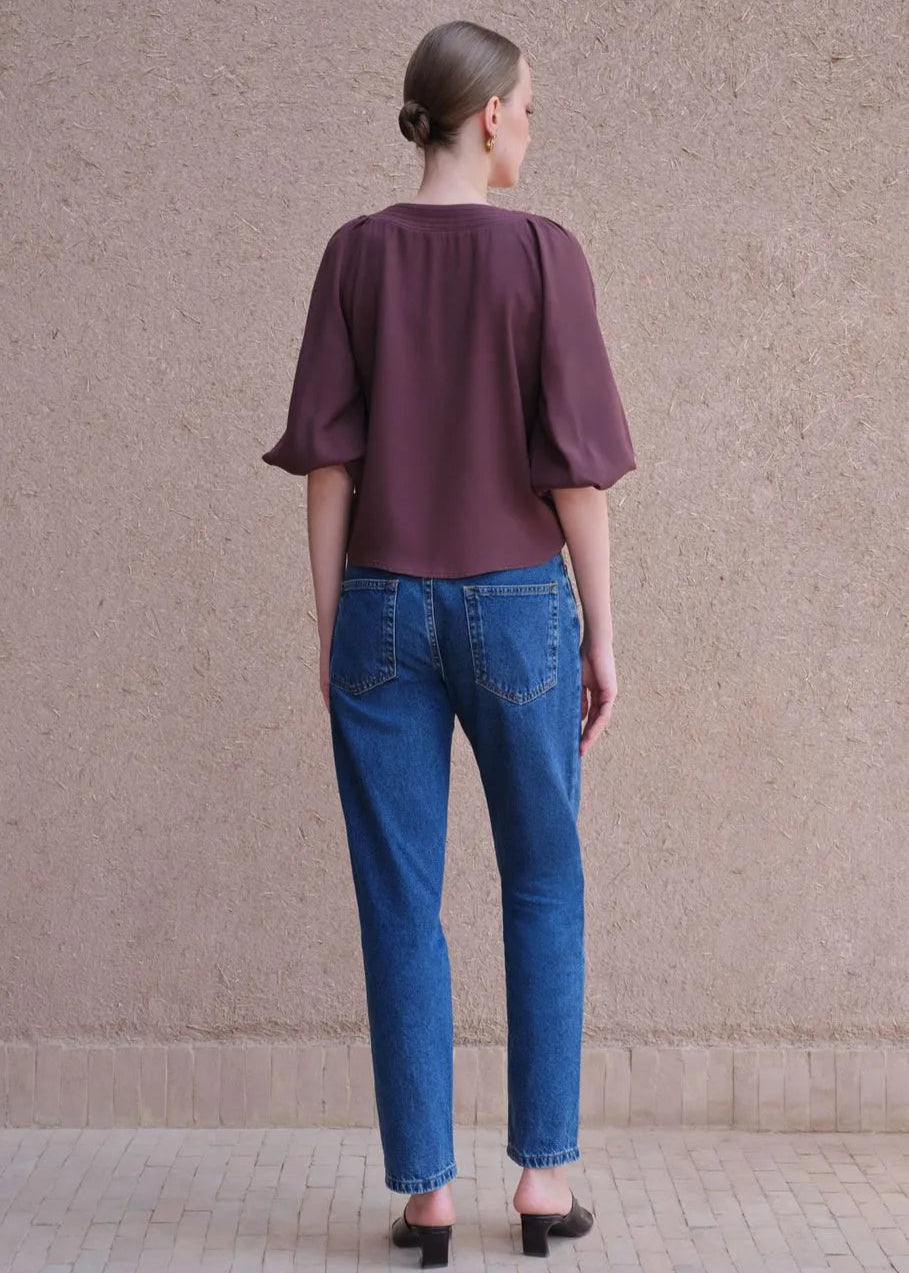 Woman wearing a purple blouse and blue jeans standing against a beige wall.