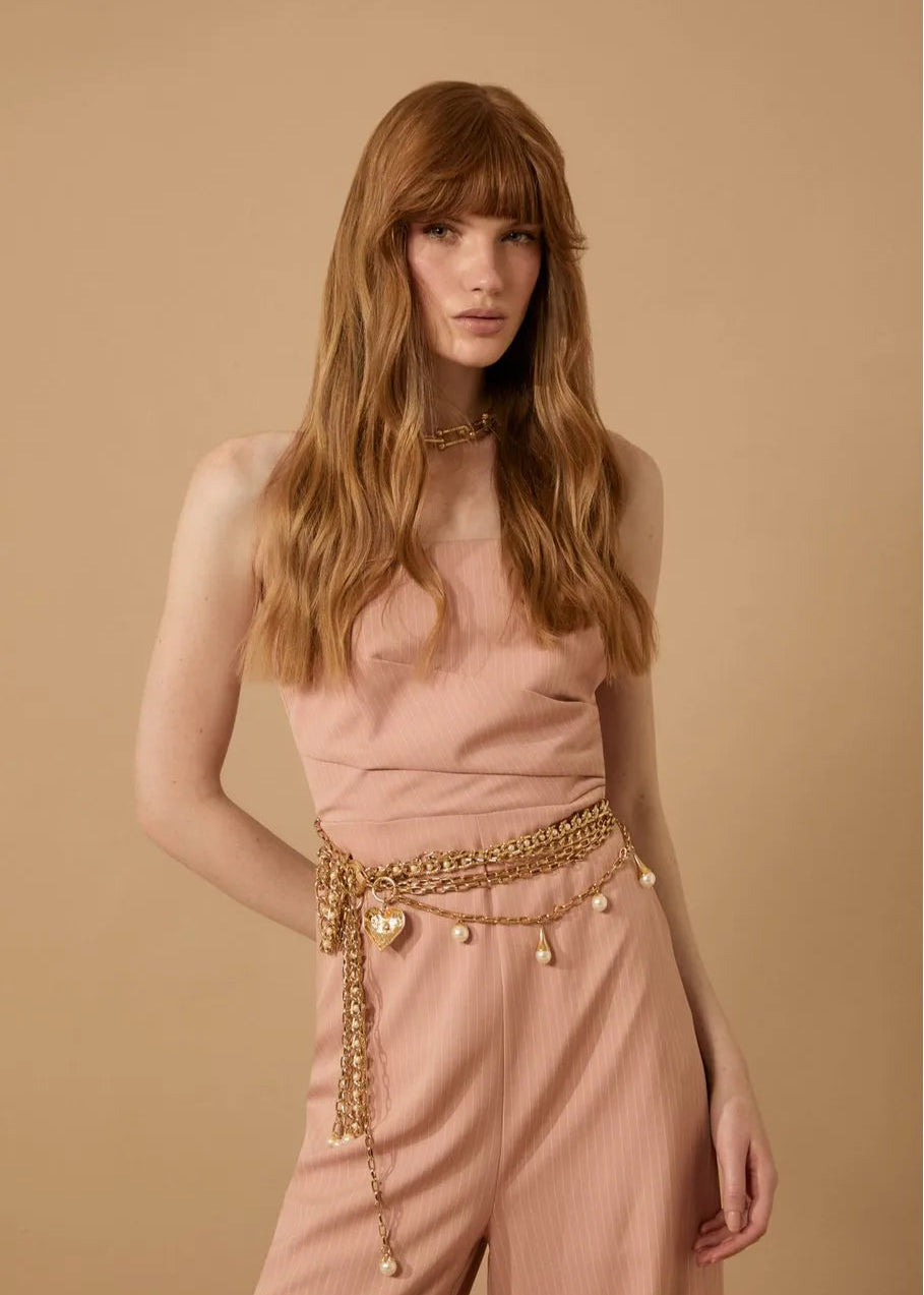 Woman wearing a pink top and pants with a gold belt against a beige background