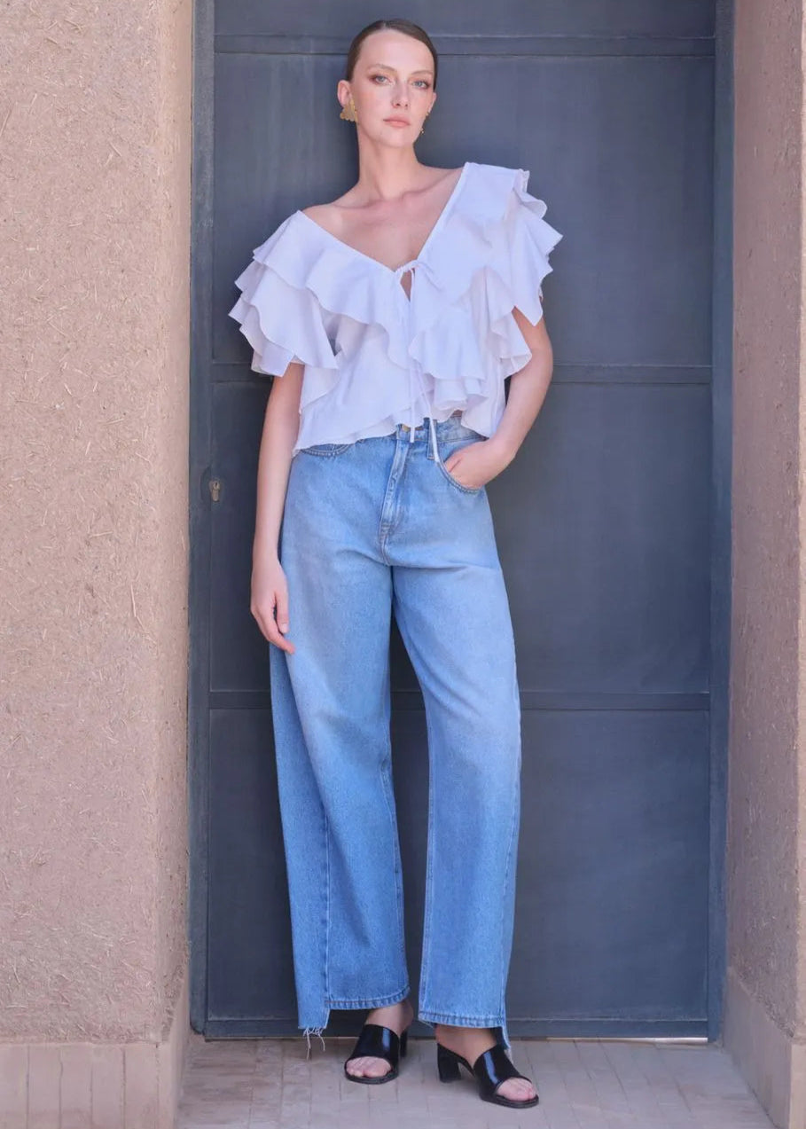 Woman wearing a white ruffled top and blue jeans standing against a plain background