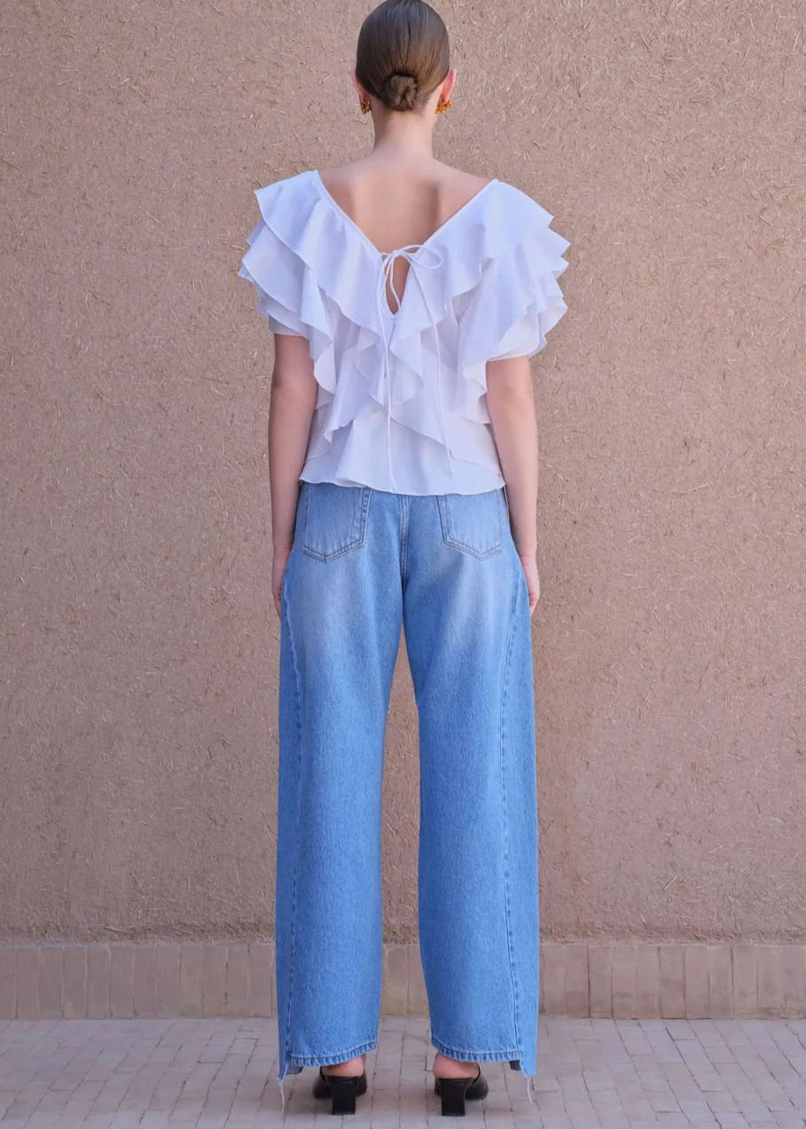 Person wearing a white ruffled blouse and blue jeans against a beige wall.