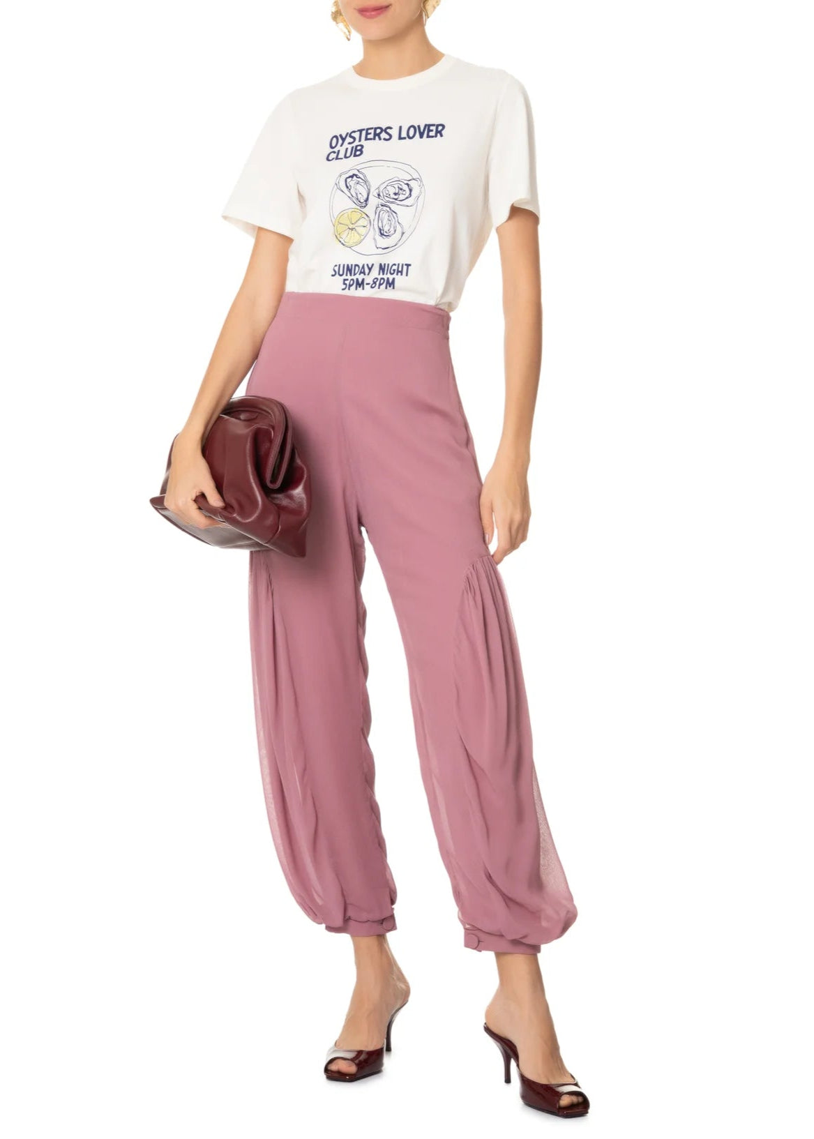 Woman wearing a white graphic t-shirt and pink pants holding a brown handbag on a white background