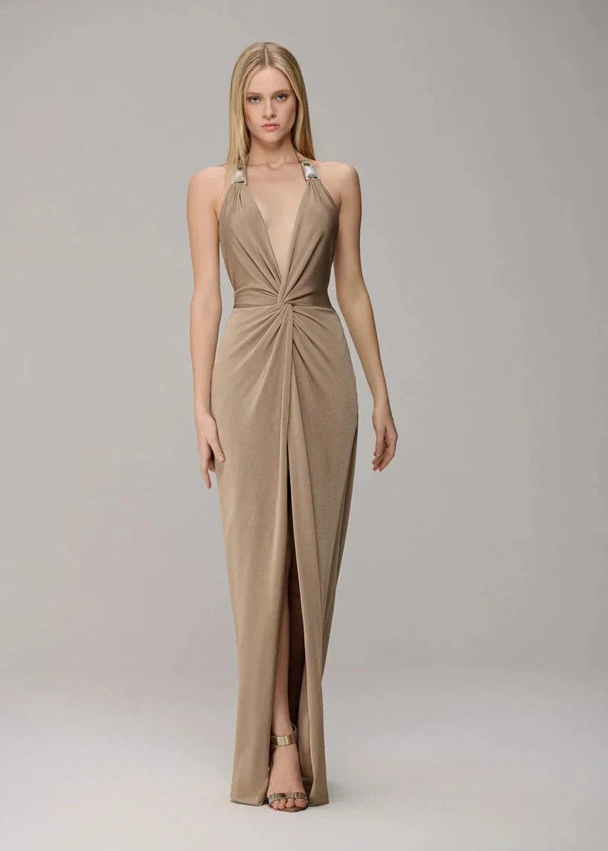 A woman modeling a floor-length halter gown in a warm taupe-beige shade against a neutral light gray studio background.