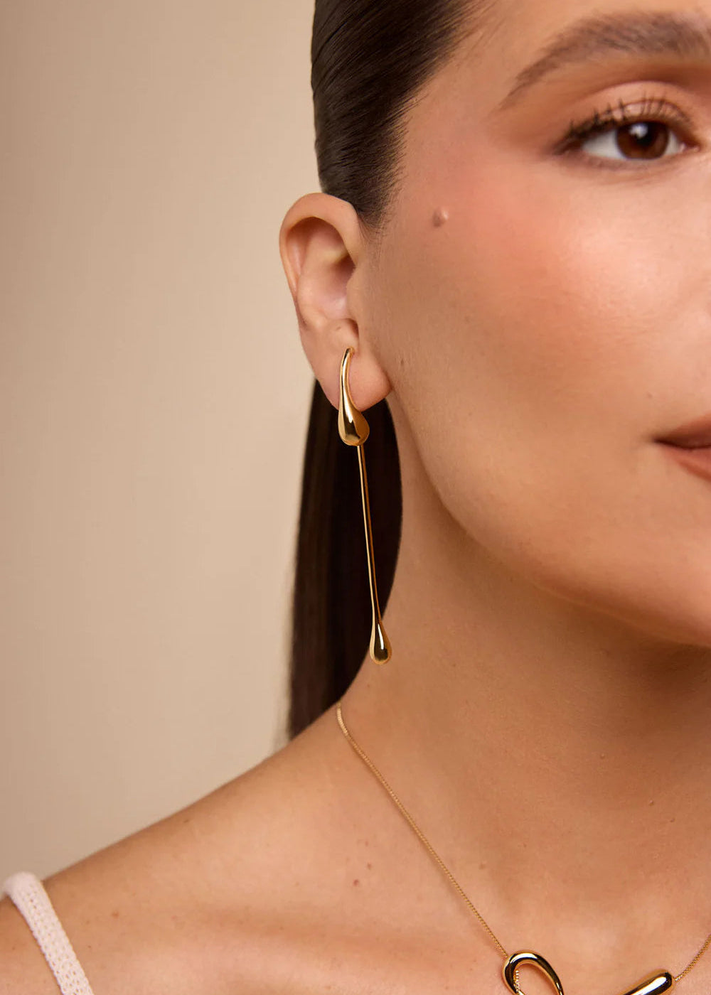 Close-up of a woman wearing gold earrings and a necklace against a beige background