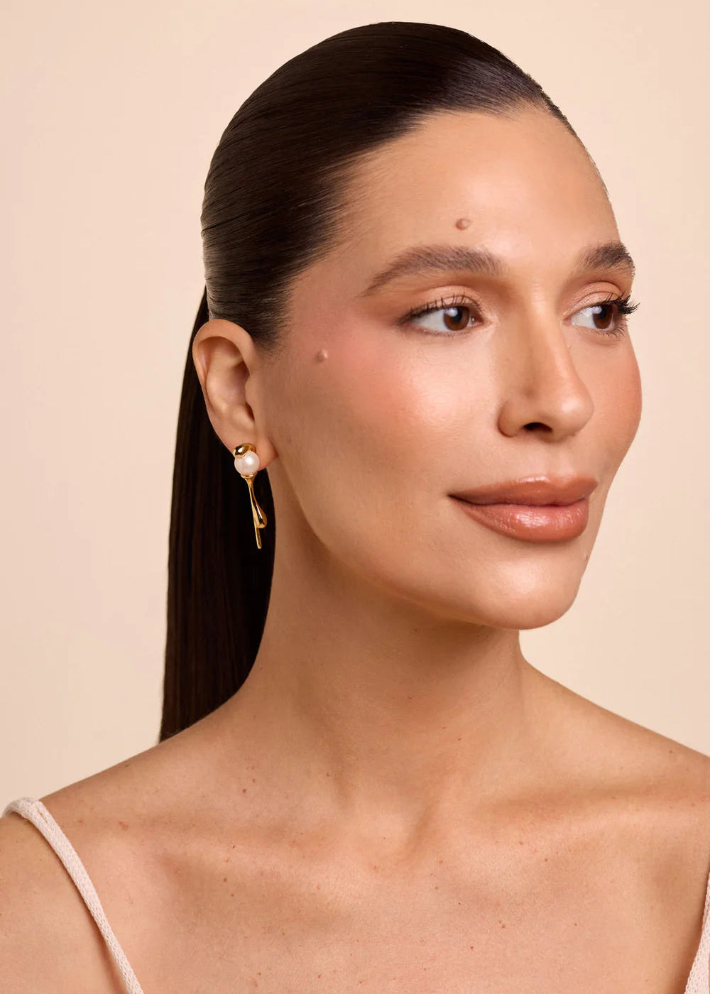 Woman with a ponytail wearing gold earrings against a beige background