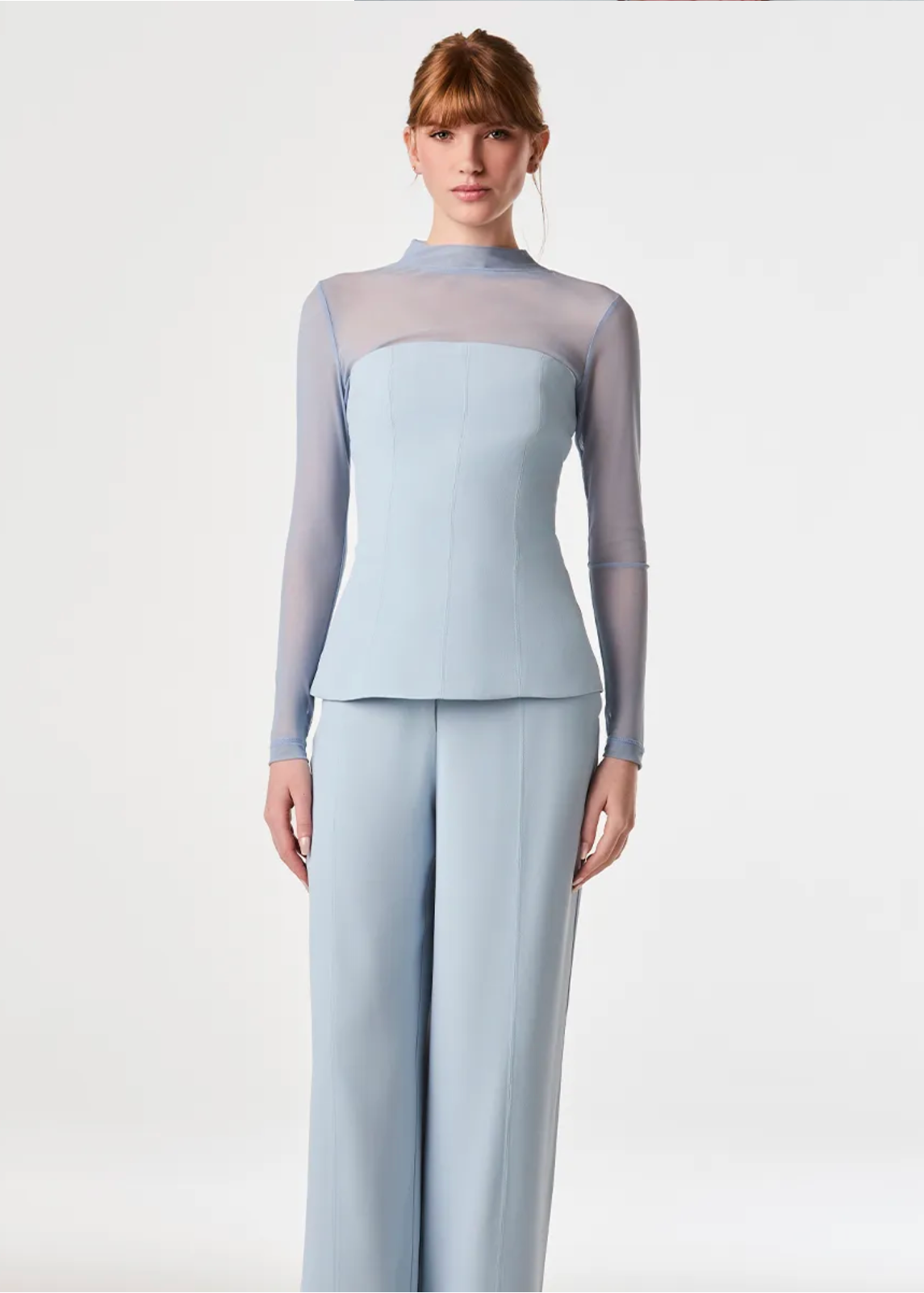 A model wearing a light blue two-piece set consisting of a mock-neck long-sleeve top with sheer mesh shoulders and matching wide-leg trousers, shot against a plain white studio background.
