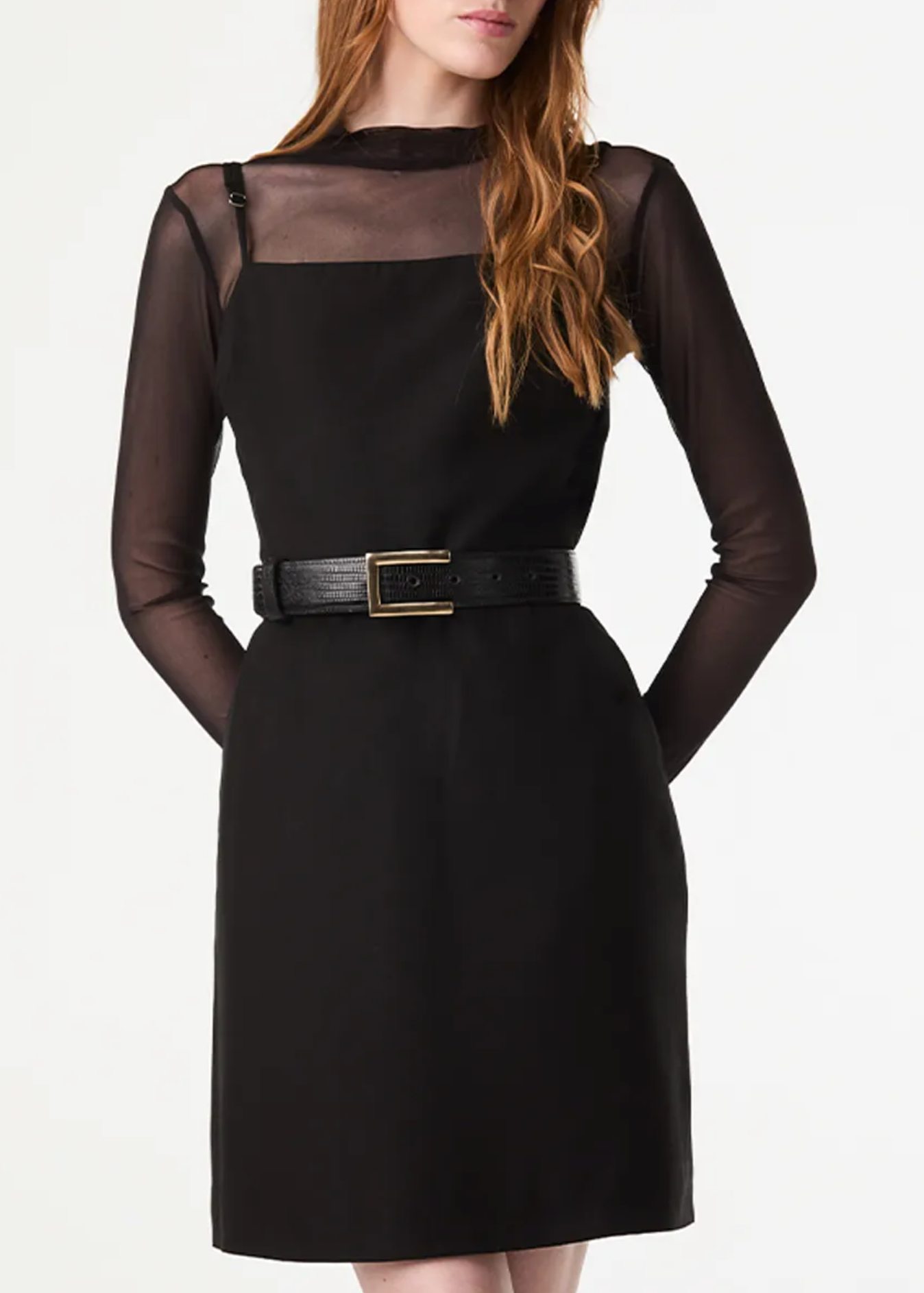 A woman wearing a black long-sleeve midi dress with sheer mesh shoulder panels, a black belt with a gold buckle, and a black collar, photographed against a plain white studio background.