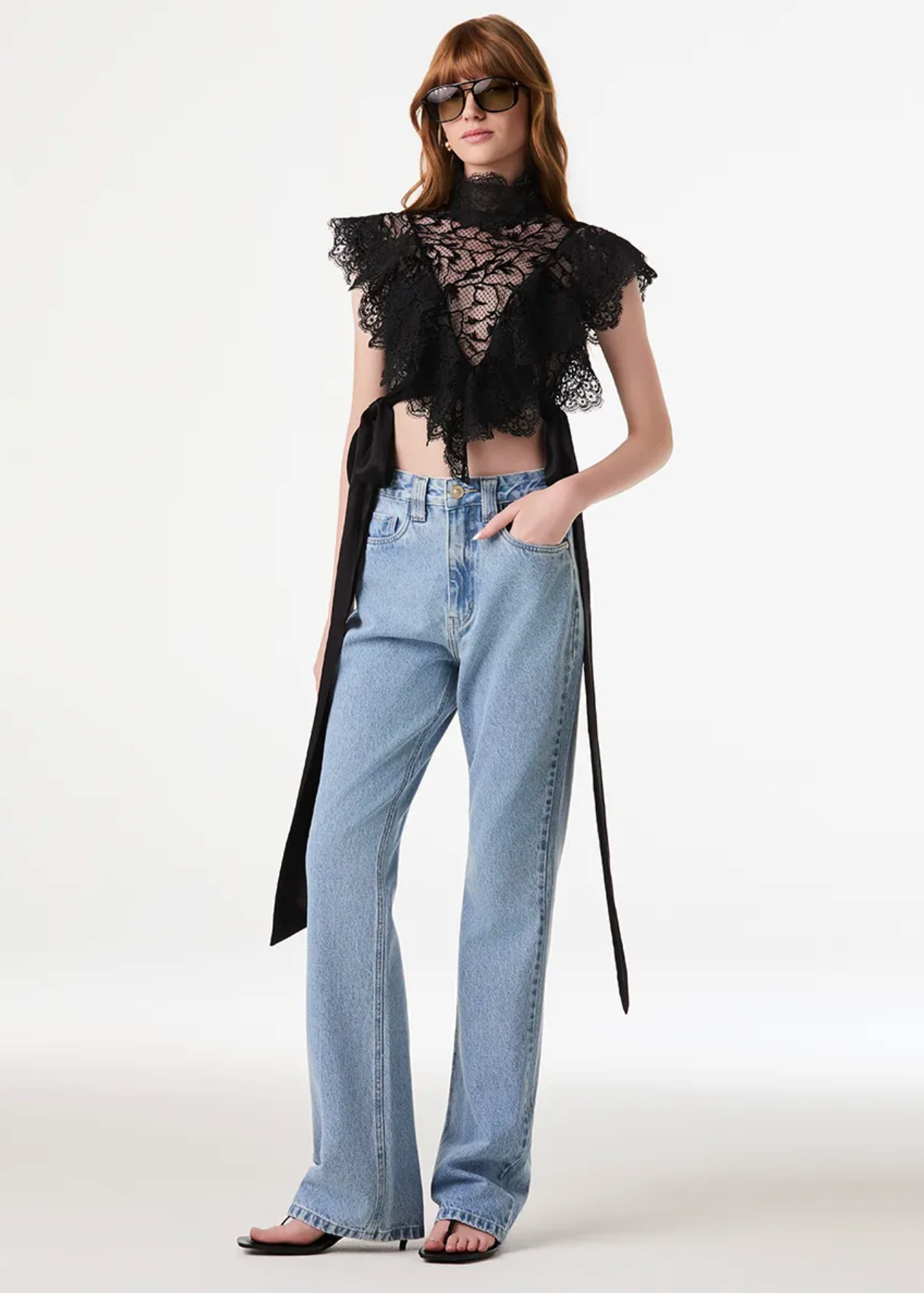 A model wearing a black ruffled sheer lace top with a long tie detail, paired with light-wash wide-leg jeans and black sandals, photographed against a plain light gray studio background.
