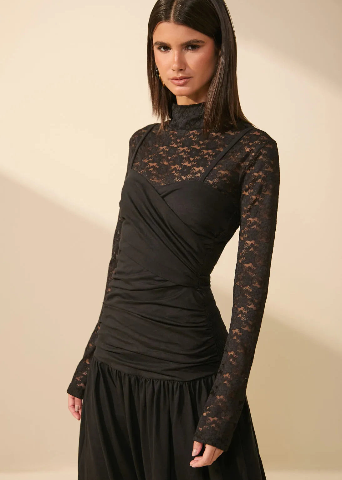 A woman modeling a black long-sleeve dress with a sheer lace mock neck and upper bodice, paired with a black skirt, against a warm beige studio background.