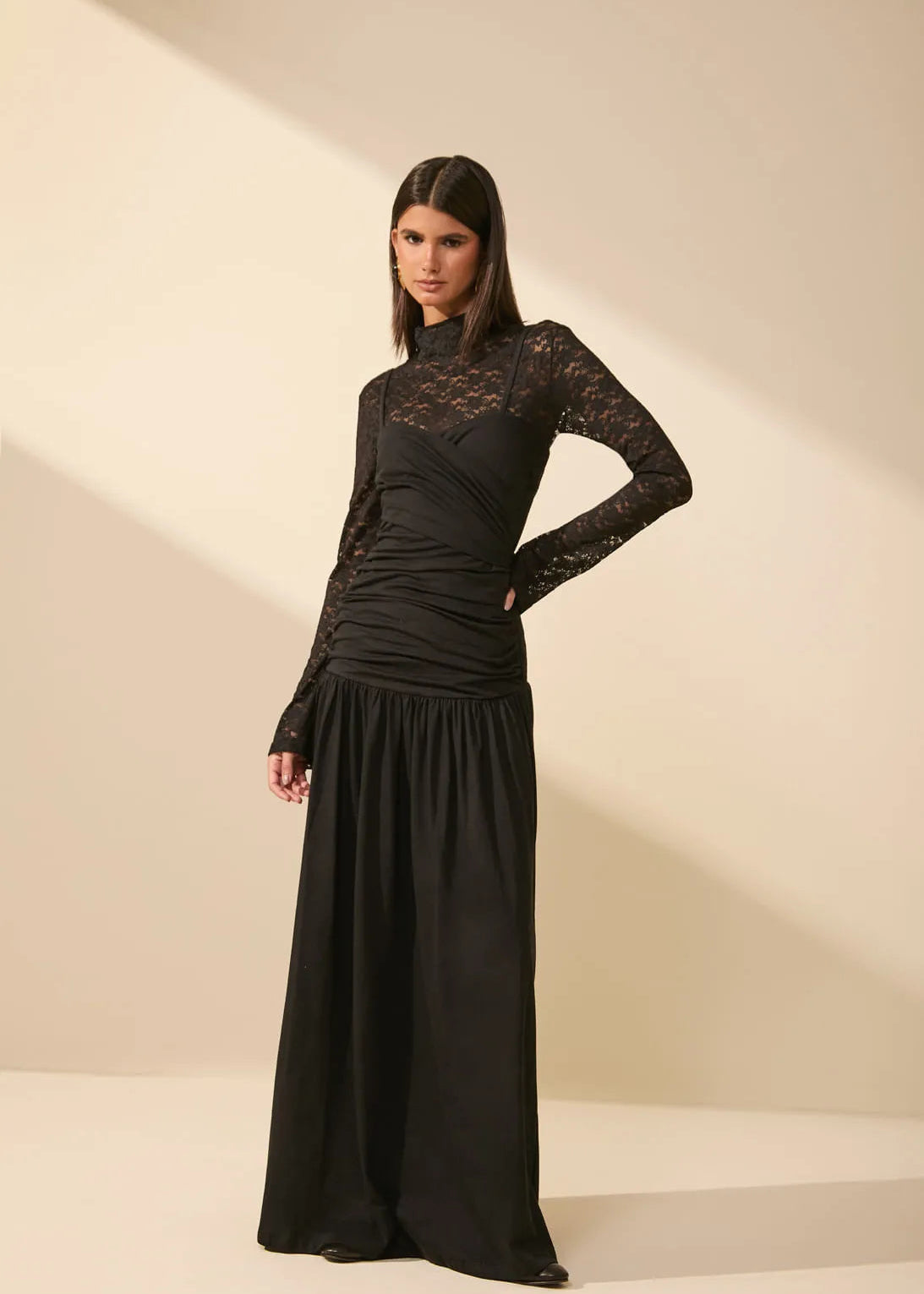 A woman modeling a long black dress with a sheer lace mock neck and long sleeves, standing against a warm beige studio backdrop with soft diagonal shadows.