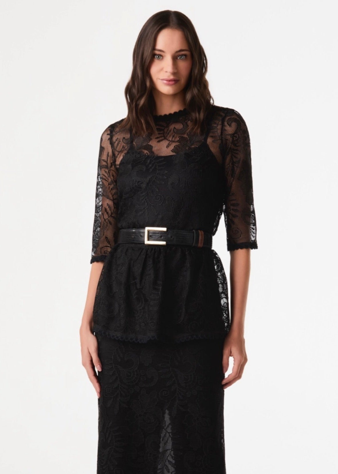 A woman modeling a black lace dress with a high mock neck, sheer three-quarter sleeves, a peplum waist, and a matching black belt with a silver buckle, set against a plain white studio background.