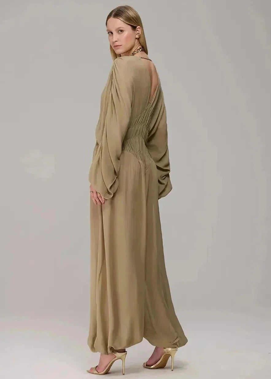A woman modeling a long, flowing maxi dress in a warm taupe-beige color, shown from a three-quarters back view against a neutral light gray studio background.