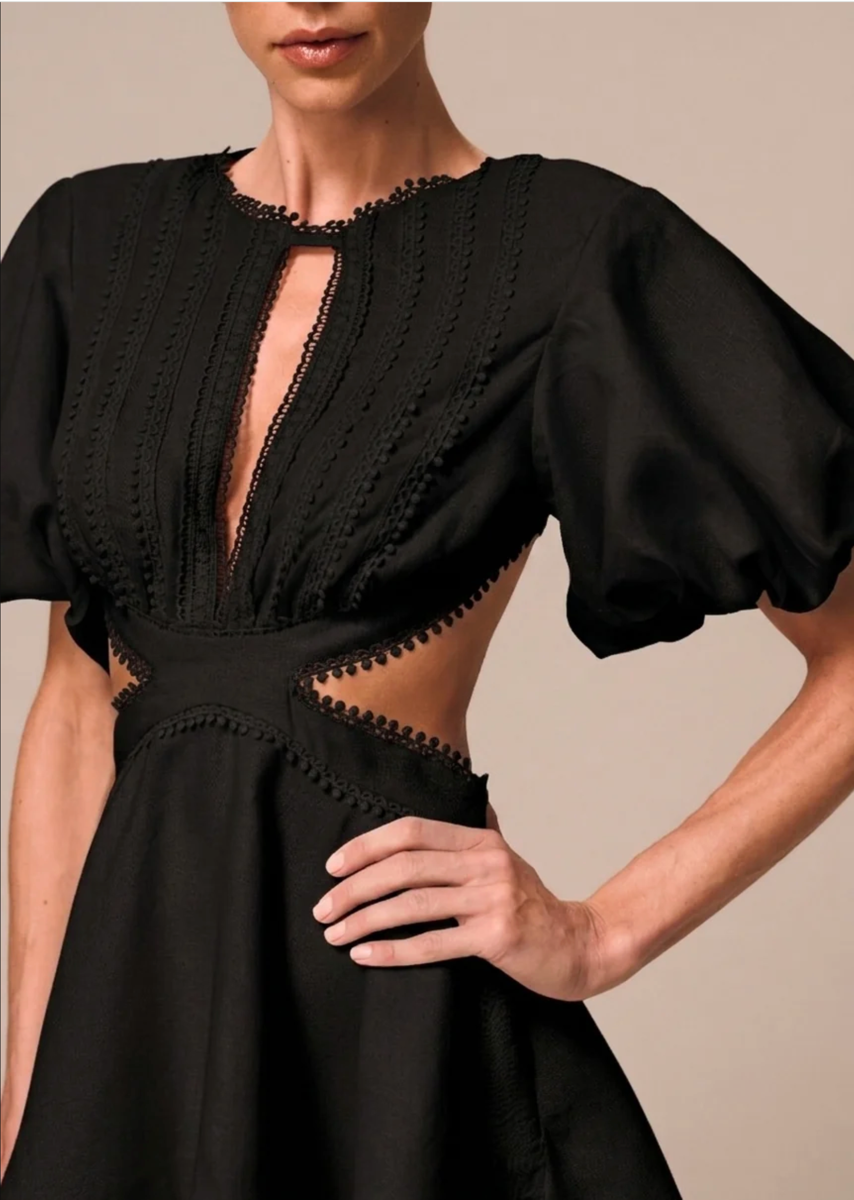 A close-up of a model wearing a black dress with dramatic puff sleeves, a deep V-neckline with a keyhole cutout, and intricate embroidered detailing around the neckline and waist.