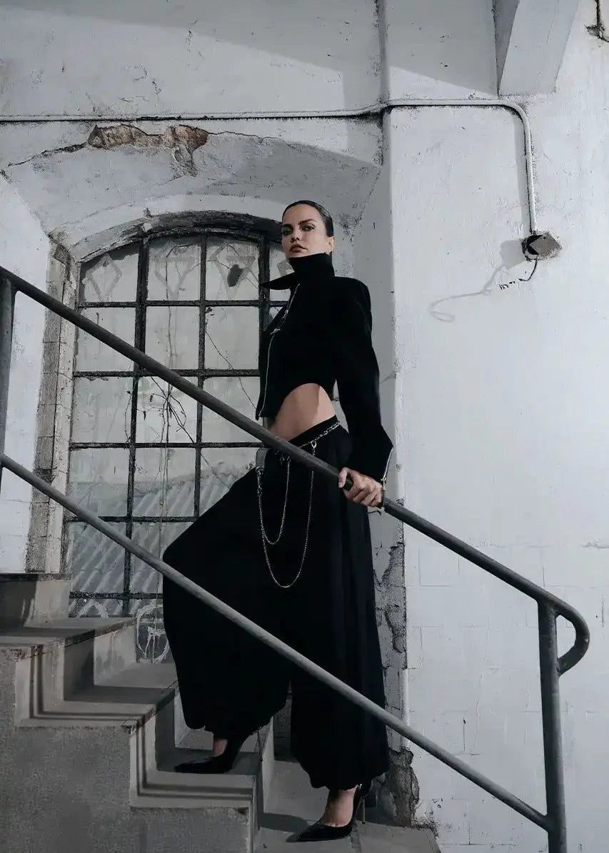 A model wearing a black high-neck long-sleeve crop top and matching wide-leg pants with silver chain details, posing on an industrial concrete staircase with a metal railing.