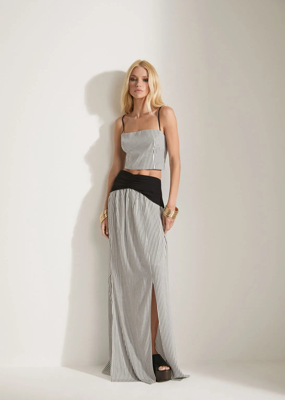 A blonde model stands against a white studio wall wearing a two-piece outfit consisting of a light gray pleated crop top with thin black straps and matching pleated wide-leg pants with a black waistband.
