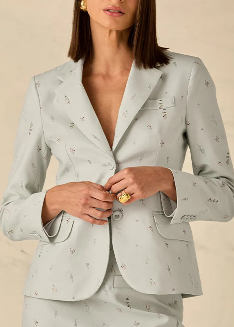 A woman wearing a light sage green blazer with a delicate floral pattern, buttoned up to the chest, set against a neutral beige background.
