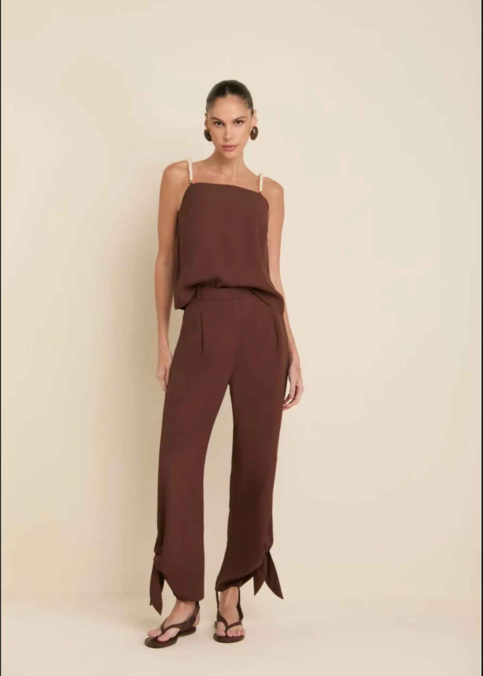 A woman modeling a brown sleeveless jumpsuit with thin shoulder straps, a draped top section, and wide-leg pants with side slits, standing against a plain beige studio background.