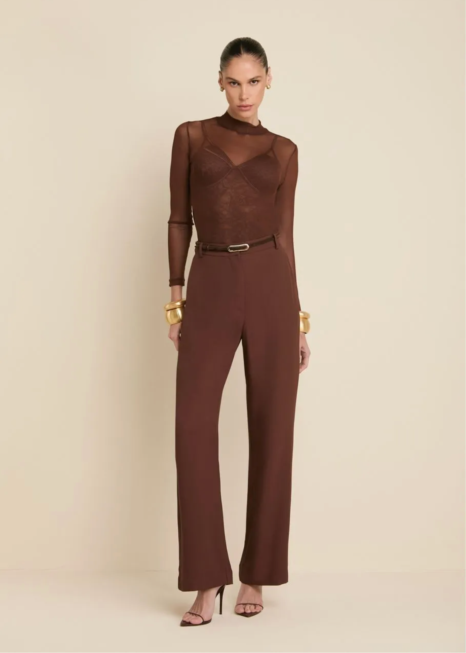 A model stands against a warm beige studio background wearing a dark brown sheer long-sleeve top with a structured bust and matching wide-leg trousers, styled with a thin belt and gold jewelry.