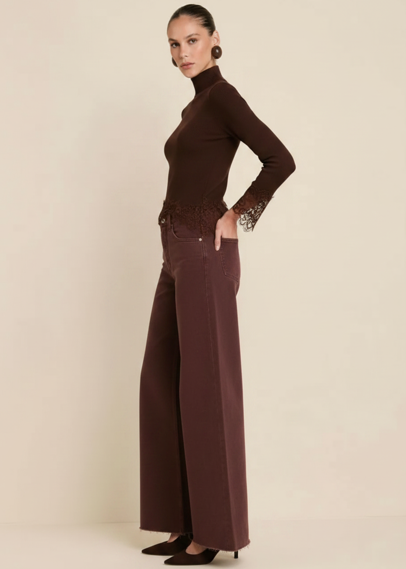 A model stands against a plain beige studio background wearing a dark brown long-sleeve mock-neck top with lace detailing at the waist and cuffs, paired with matching wide-leg trousers and black pointed heels.
