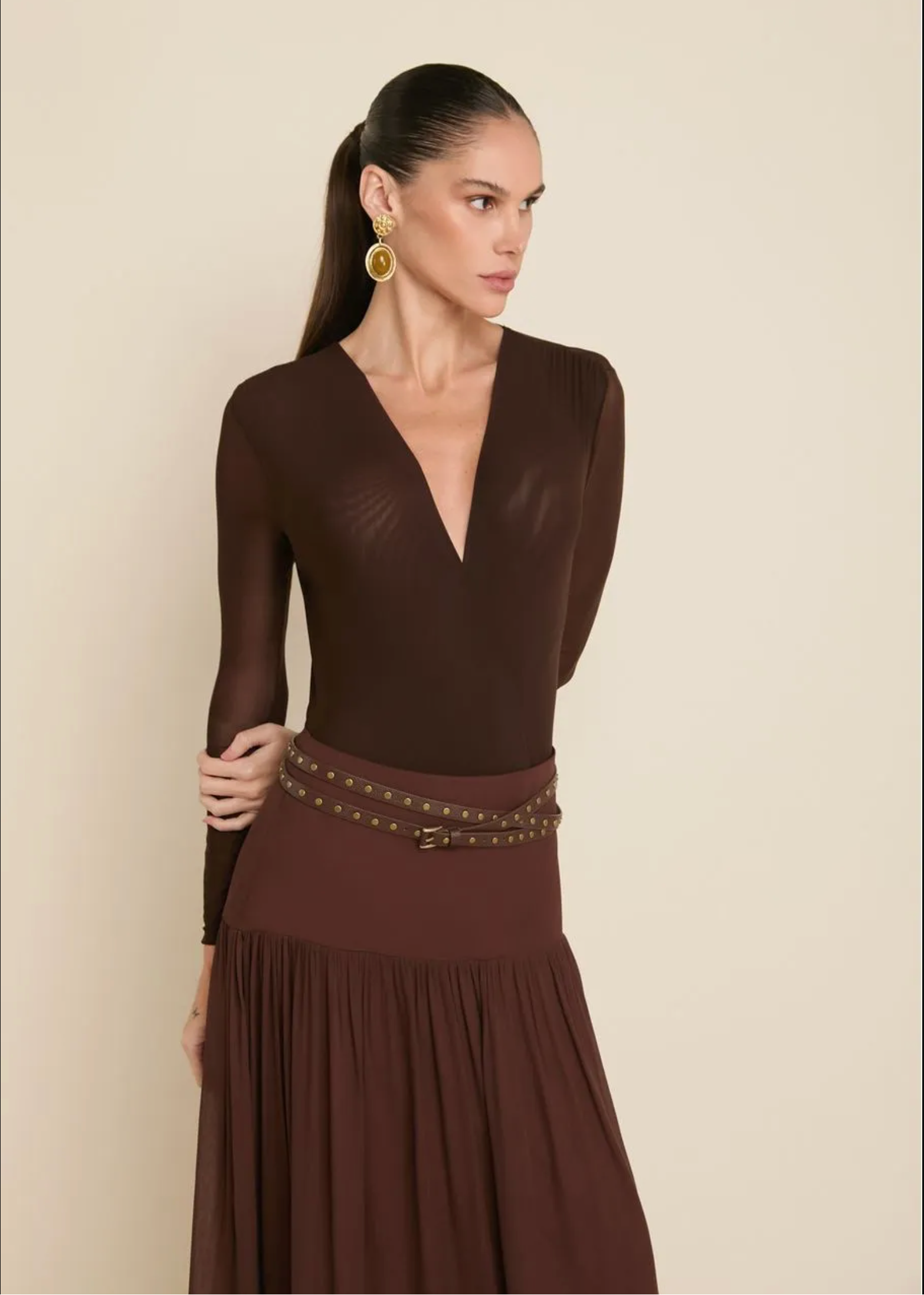 A woman modeling a dark brown long-sleeve dress with a deep V-neckline, sheer upper bodice, and a pleated skirt, accessorized with a studded belt. She is wearing large gold hoop earrings against a plain beige background.