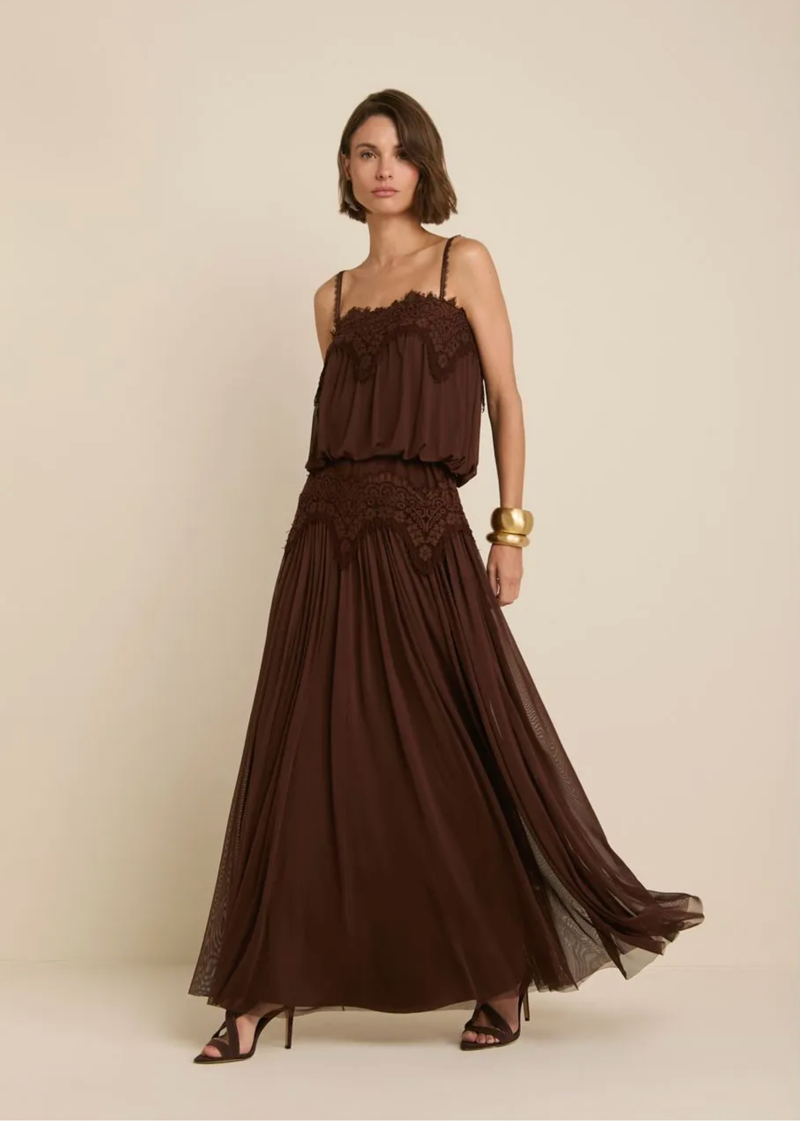 A woman modeling a dark brown sleeveless maxi dress with a ruffled bodice, lace waistband, and flowing pleated skirt, standing against a plain beige studio background.