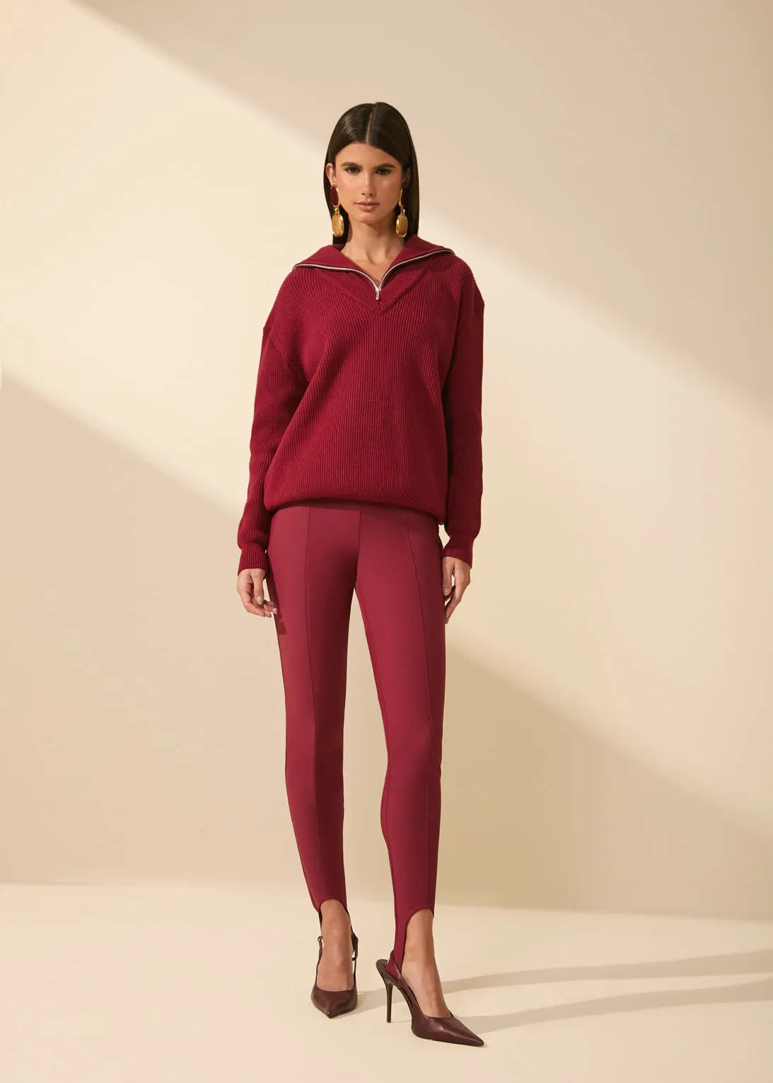 A model stands in a studio wearing a matching burgundy knit half-zip pullover and leggings set, paired with dark brown pointed-toe heels, against a warm beige background with soft diagonal shadows.