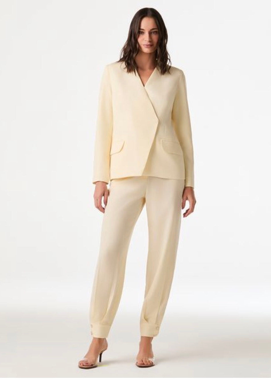A woman modeling a light cream-colored two-piece suit consisting of a wrap-style blazer and matching slim-fit trousers, paired with clear high-heeled sandals against a plain white studio background.