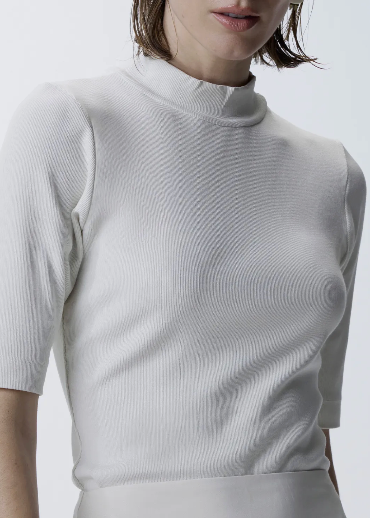 A close-up of a model wearing a white ribbed short-sleeve mock neck top against a light gray studio background.
