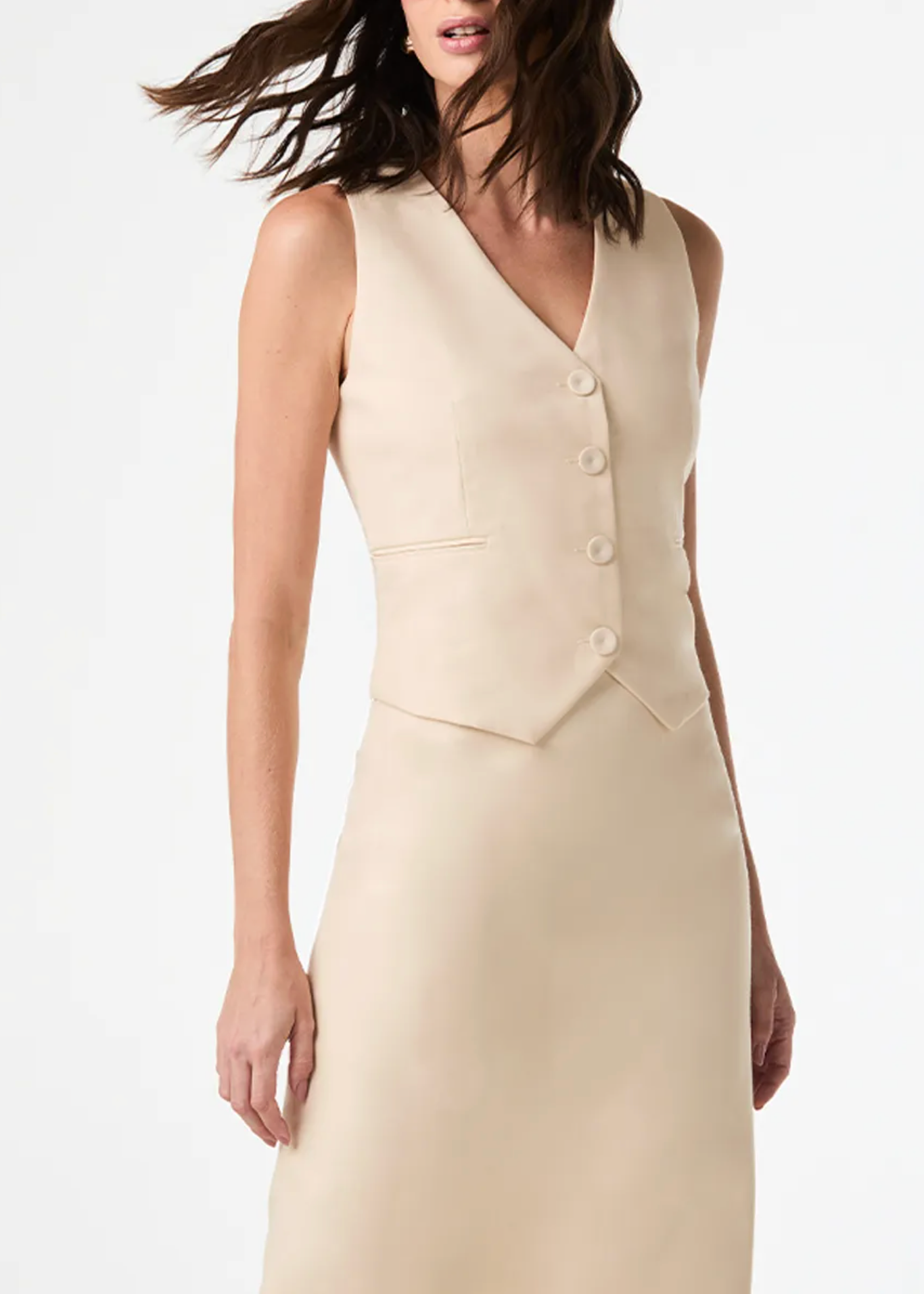 A woman wearing a sleeveless beige vest and matching midi skirt against a clean white studio background.