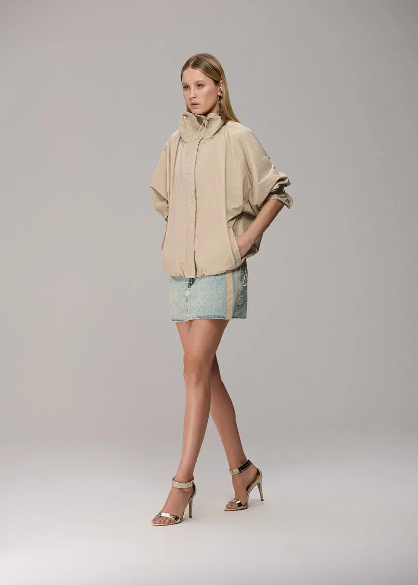 A model wearing a beige hooded jacket with a high collar, paired with a light blue denim mini skirt and beige high-heeled sandals, standing against a neutral gray studio background.