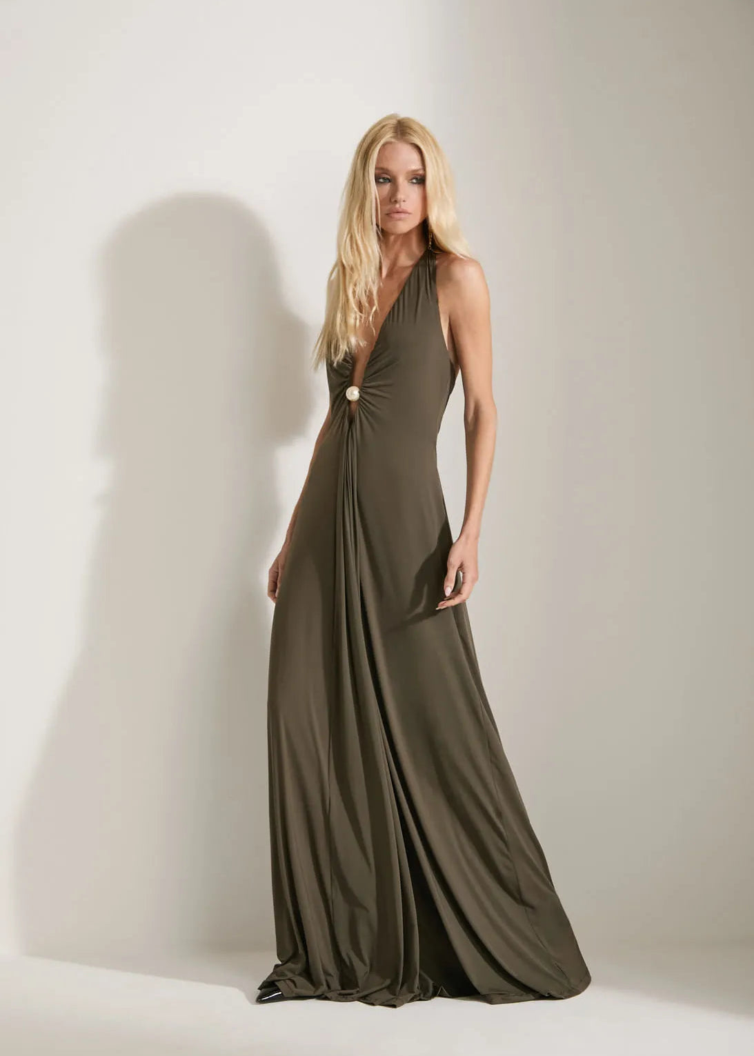 A blonde model wears a floor-length, olive green halter gown with a gathered waist detail, standing against a neutral light gray studio background.