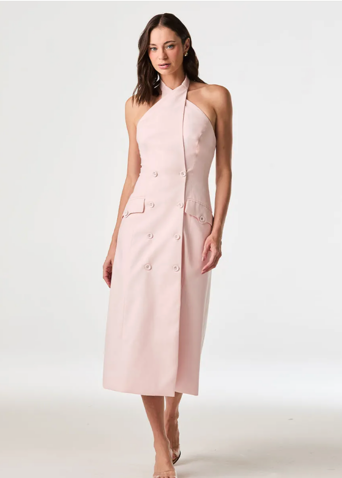 A woman modeling a sleeveless, halter-neck midi dress in a soft light pink. The dress features a structured double-breasted front with decorative buttons and flap pockets on the sides, photographed against a seamless light gray studio background.