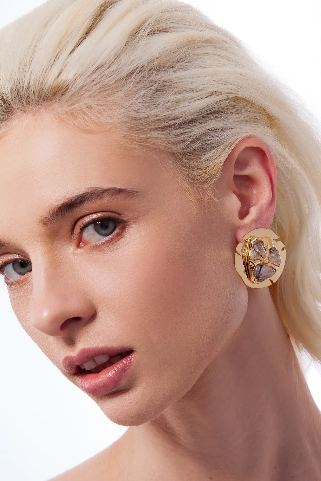 A close-up of a blonde woman wearing a large gold-toned statement earring with a floral or clover-like design and clear stones, against a clean white background.