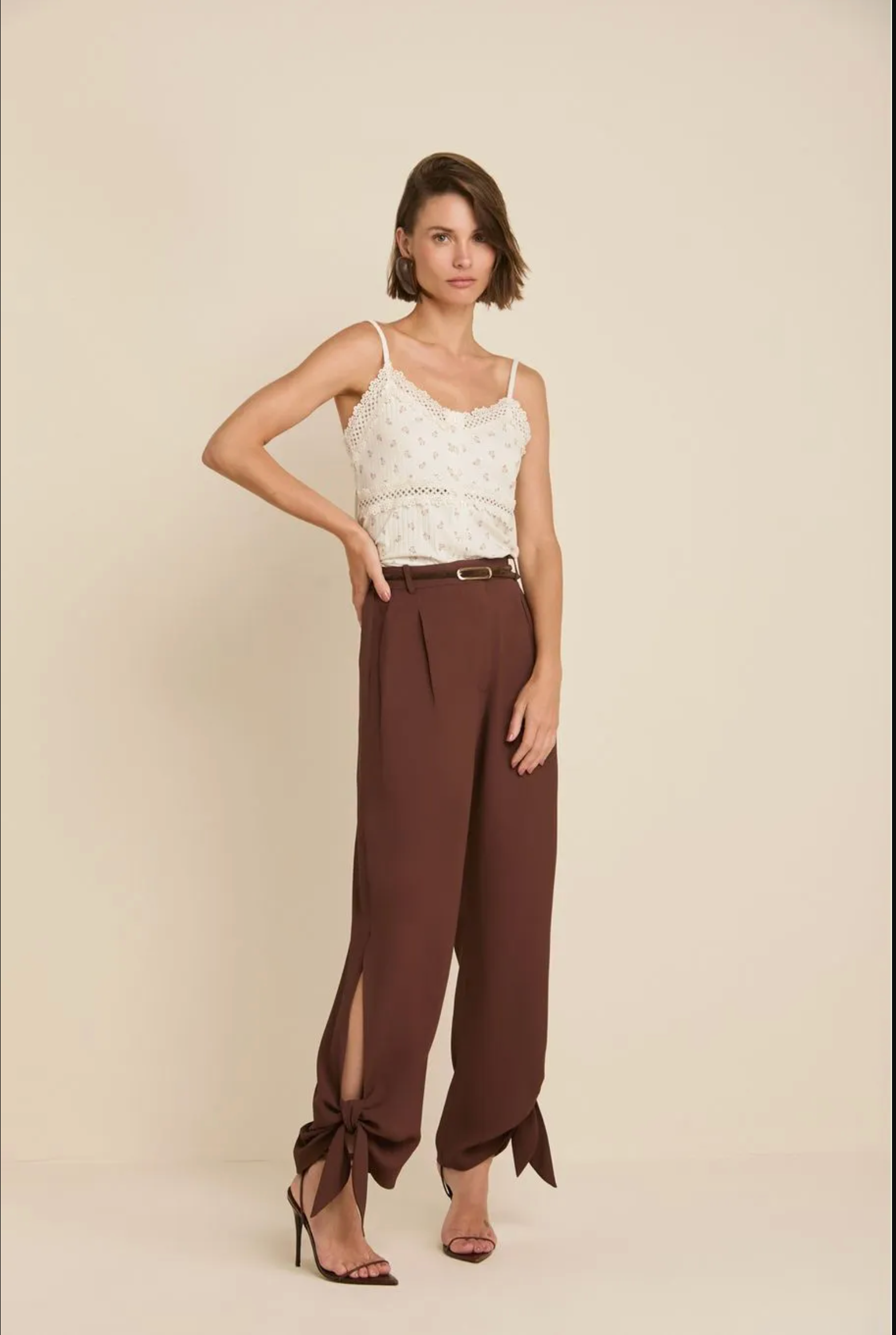 A model stands against a plain beige studio background wearing a white lace camisole top and high-waisted brown wide-leg trousers with side slits and bow details at the ankles, paired with black heeled sandals.