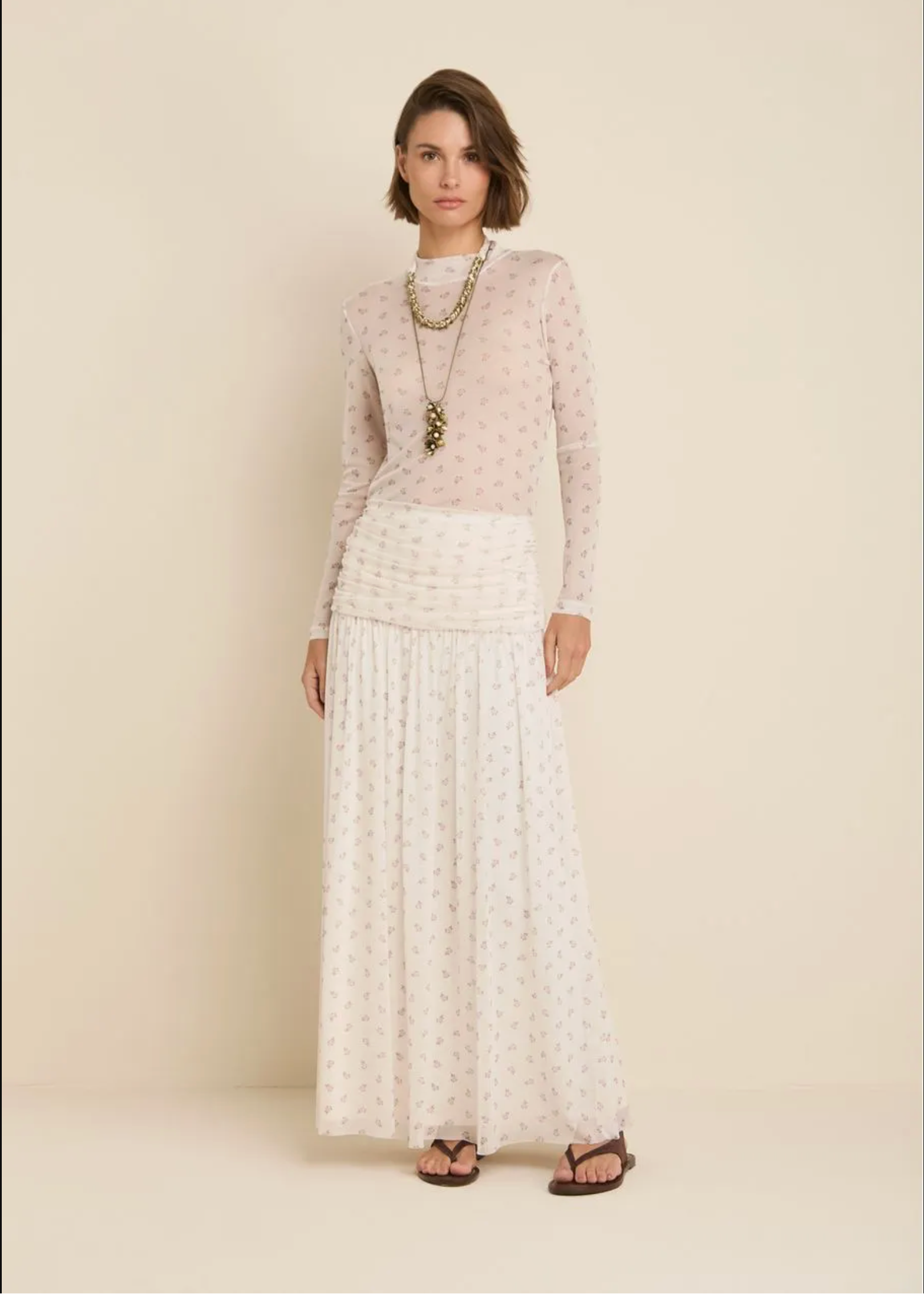 A woman stands against a plain warm beige studio background wearing a sheer long-sleeve top and matching maxi skirt set in a light cream color with a small repeating floral print. She is styled with layered gold necklaces and brown sandals.