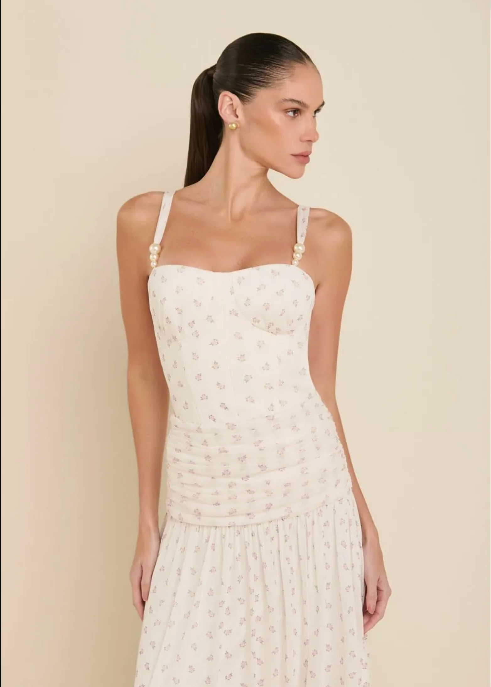 A woman modeling a white spaghetti-strap dress with a delicate floral pattern and pearl-like bead details on the straps, set against a solid beige background.