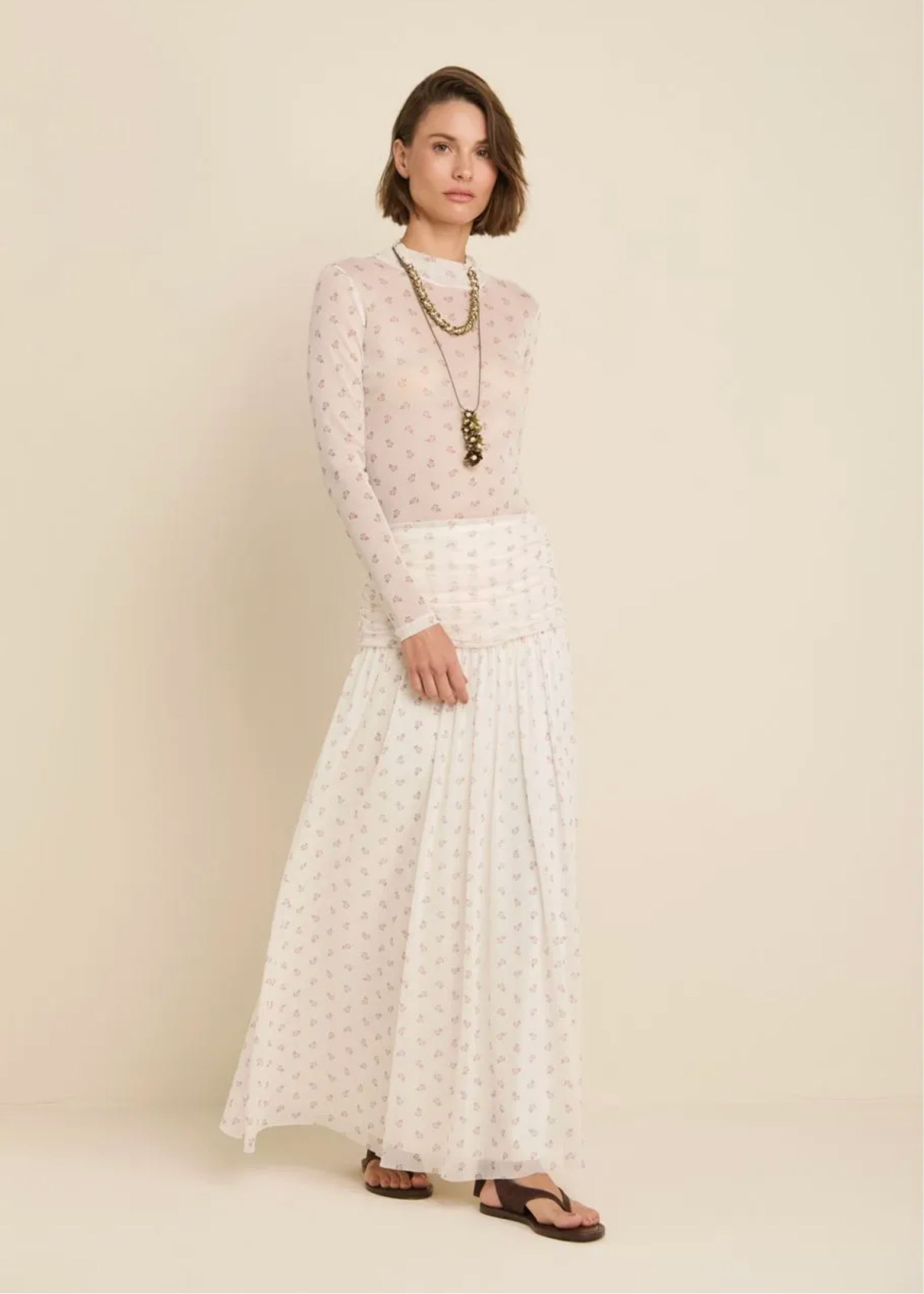 A woman stands against a plain beige studio background wearing a sheer long-sleeve top with a small repeating floral print over a white long-sleeve maxi skirt with a subtle allover print. She is wearing layered gold necklaces and brown sandals.