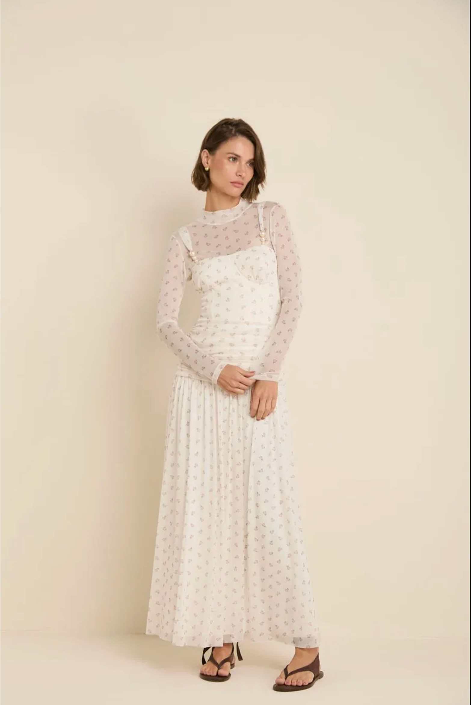 A woman modeling a long-sleeve white dress with a delicate pink floral pattern, featuring a sheer mesh mock neck overlay and a pleated skirt, set against a solid cream background.