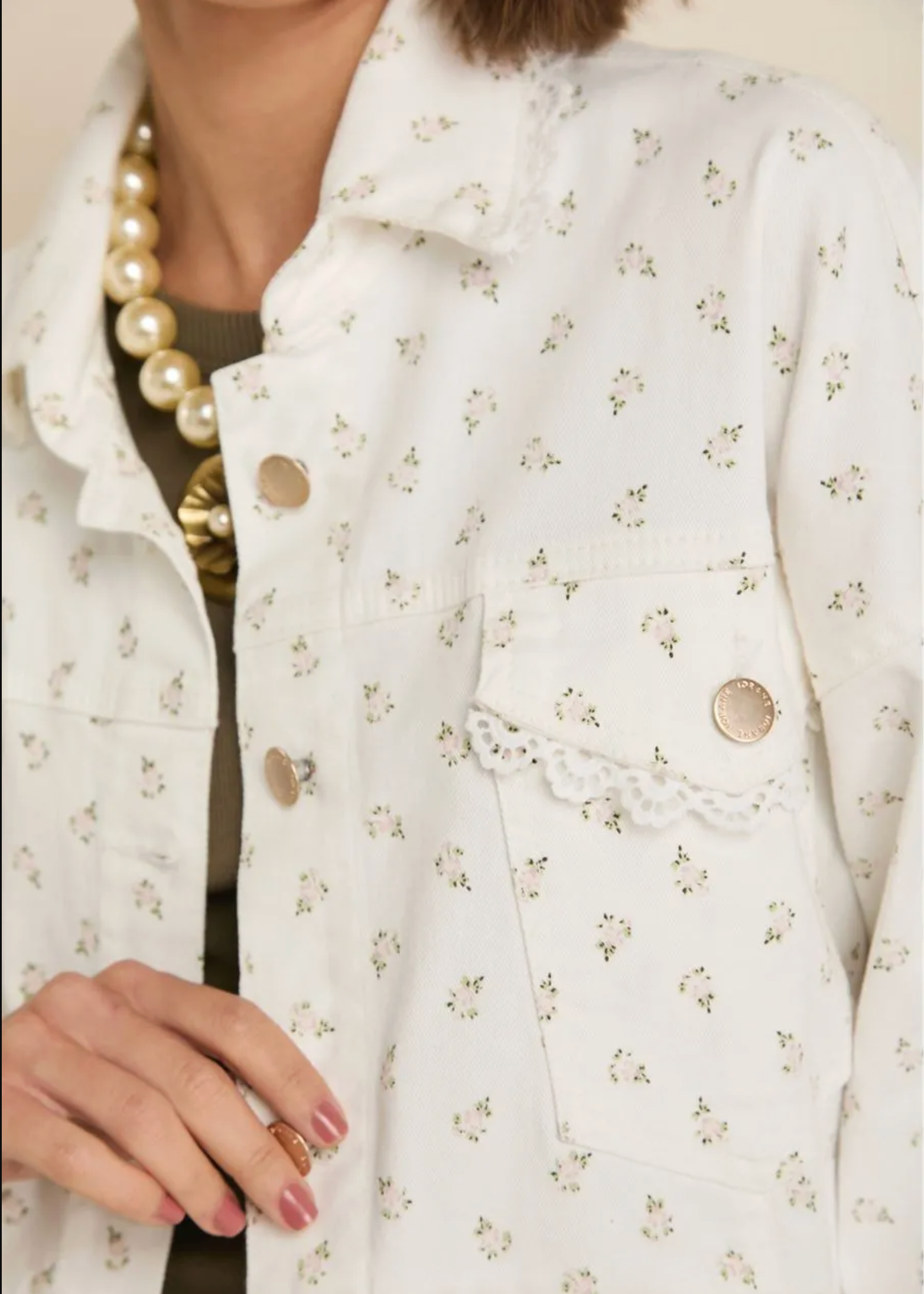 Close-up of a person wearing a white button-front jacket with a small scattered floral print, lace trim along the pocket edge, and gold-tone buttons. A pearl necklace and ring are also visible.