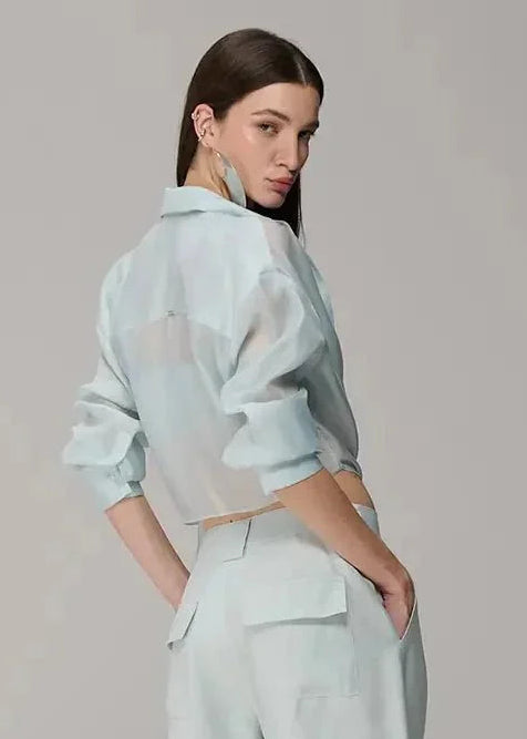 A model is shown from the back wearing a light mint green two-piece outfit consisting of a long-sleeve collared shirt and matching wide-leg trousers with sheer mesh paneling at the lower legs, set against a plain light gray studio background.