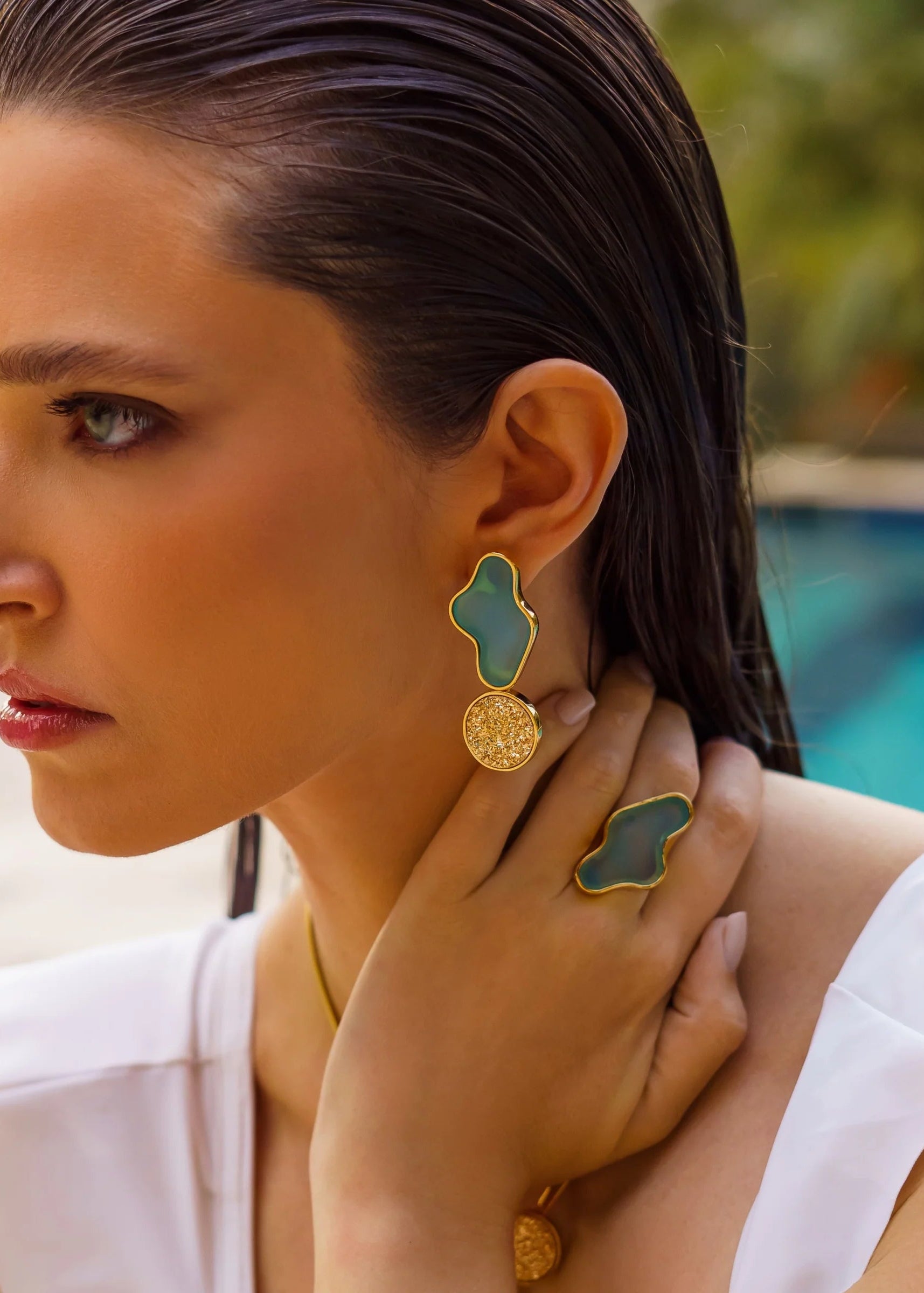 Woman wearing gold and green earrings with a blurred pool background
