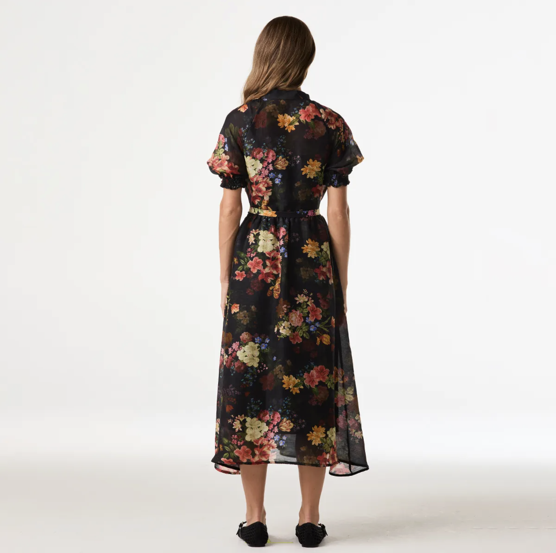 A woman viewed from the back wearing a short-sleeve midi dress with a black base and a colorful floral print, paired with black flats, against a plain white studio background.
