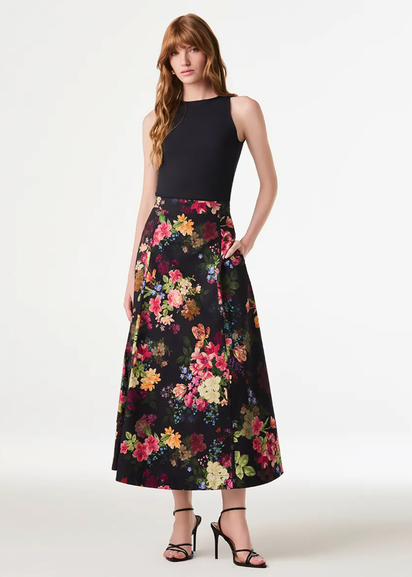 A model stands against a plain light background wearing a sleeveless black top and a high-waisted midi skirt with a dense multicolor floral print on a black base. She is wearing black strappy high-heeled sandals.